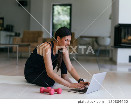A young woman in sportswear is sitting in the living room and preparing for online training while using a laptop. A young woman in sportswear is sitting in the living room and preparing for online training while using a laptop. 129884460