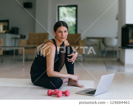 A young woman in sportswear is sitting in the living room and preparing for online training while using a laptop. 129884461