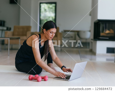 A young woman in sportswear is sitting in the living room and preparing for online training while using a laptop. A young woman in sportswear is sitting in the living room and preparing for online training while using a laptop. 129884472
