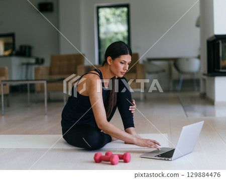 A young woman in sportswear is sitting in the living room and preparing for online training while using a laptop. 129884476