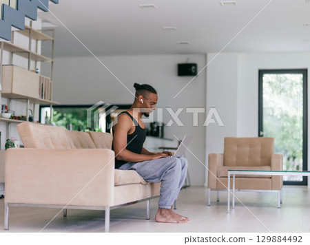 African American man in glasses sitting at a table in a modern living room, using a laptop for business video chat, conversation with friends and entertainment. 129884492