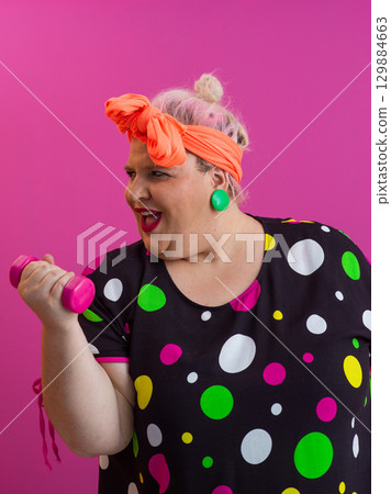 Surprised plus size woman exercising with pink dumbbells on colorful background 129884663