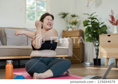 Confident Overweight Woman Exercising at Home with Yoga Mat and Laptop in Bright Living Room Setting 129884879