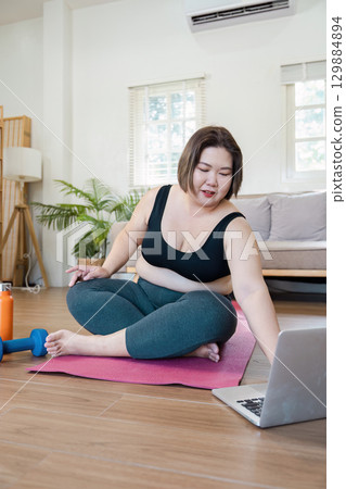Overweight Woman Exercising at Home with Yoga Mat and Laptop for Online Fitness Class 129884894