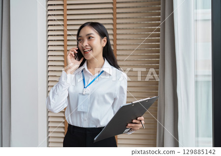 Confident Business Professional Engaged in a Phone Call While Holding a Clipboard in a Modern Office Setting 129885142