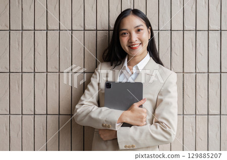 Confident Businesswoman Holding Tablet Against Modern Textured Wall in Professional Attire 129885207
