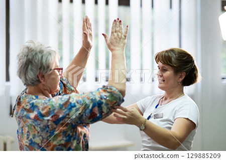 Senior Physical Therapy Session.Senior woman doing physical exercises with the help of a smiling therapist. Concept of elderly rehabilitation and movement recovery. Senior Physical Therapy Session.Senior woman doing physical exercises with the help of a smiling therapist. Concept of elderly rehabilitation and movement recovery. 129885209