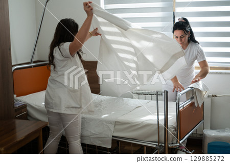 Two nurses in uniforms cleaning and organizing a hospital room by changing the bed linens and ensuring a hygienic environment for patients. Two nurses in uniforms cleaning and organizing a hospital room by changing the bed linens and ensuring a hygienic environment for patients. 129885272