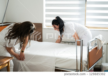 Two nurses in uniforms cleaning and organizing a hospital room by changing the bed linens and ensuring a hygienic environment for patients. 129885302
