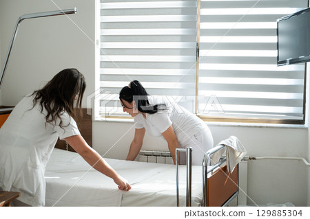 Two nurses in uniforms cleaning and organizing a hospital room by changing the bed linens and ensuring a hygienic environment for patients. 129885304