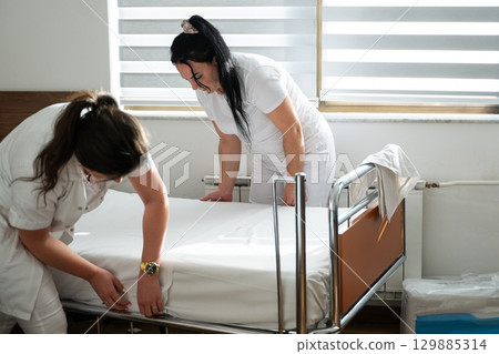 Two nurses in uniforms cleaning and organizing a hospital room by changing the bed linens and ensuring a hygienic environment for patients. Two nurses in uniforms cleaning and organizing a hospital room by changing the bed linens and ensuring a hygienic environment for patients. 129885314