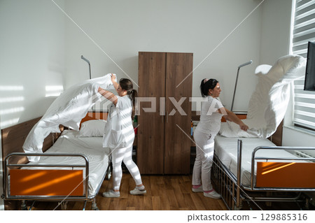 Two nurses in uniforms cleaning and organizing a hospital room by changing the bed linens and ensuring a hygienic environment for patients. Two nurses in uniforms cleaning and organizing a hospital room by changing the bed linens and ensuring a hygienic environment for patients. 129885316