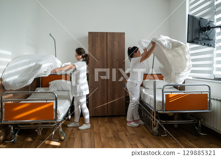 Two nurses in uniforms cleaning and organizing a hospital room by changing the bed linens and ensuring a hygienic environment for patients. 129885321