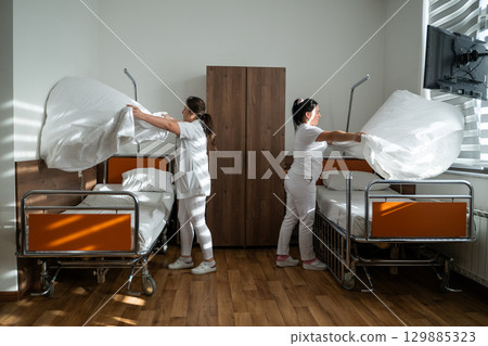 Two nurses in uniforms cleaning and organizing a hospital room by changing the bed linens and ensuring a hygienic environment for patients. Two nurses in uniforms cleaning and organizing a hospital room by changing the bed linens and ensuring a hygienic environment for patients. 129885323