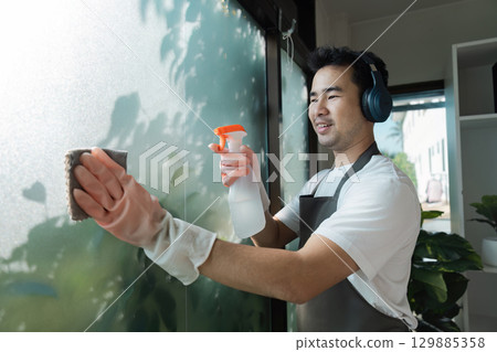 Man Cleaning Window with Spray Bottle and Sponge Wearing Headphones in Modern Home Environment 129885358