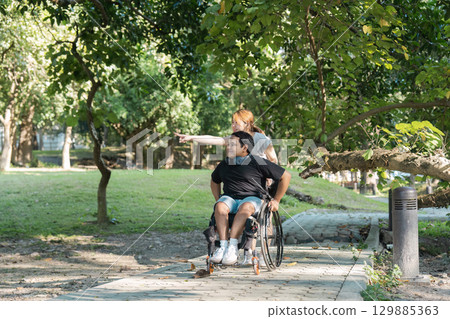 Inclusive Outdoor Activity: Young Man in Wheelchair Enjoying Nature with Friend in Park Setting 129885363