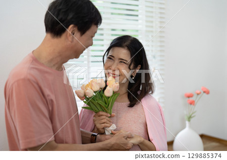 Happy Senior Couple Celebrating Valentine's Day with Flowers and Smiles in Cozy Home Setting Happy Senior Couple Celebrating Valentine's Day with Flowers and Smiles in Cozy Home Setting 129885374