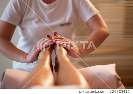Foot Massage Therapy in a Relaxing Spa Setting. A professional massage therapist in a white uniform provides a soothing foot massage to a client in a calm and serene spa environment 129885415