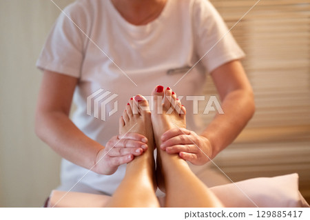 Foot Massage Therapy in a Relaxing Spa Setting. A professional massage therapist in a white uniform provides a soothing foot massage to a client in a calm and serene spa environment 129885417