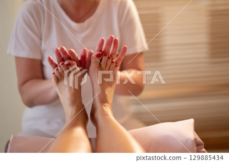 Foot Massage Therapy in a Relaxing Spa Setting. A professional massage therapist in a white uniform provides a soothing foot massage to a client in a calm and serene spa environment 129885434