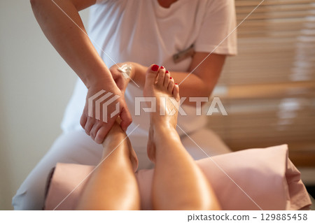 Foot Massage Therapy in a Relaxing Spa Setting. A professional massage therapist in a white uniform provides a soothing foot massage to a client in a calm and serene spa environment 129885458