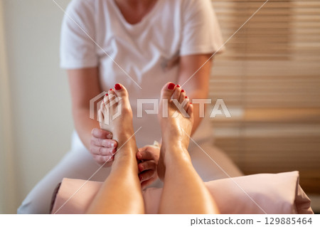 Foot Massage Therapy in a Relaxing Spa Setting. A professional massage therapist in a white uniform provides a soothing foot massage to a client in a calm and serene spa environment 129885464