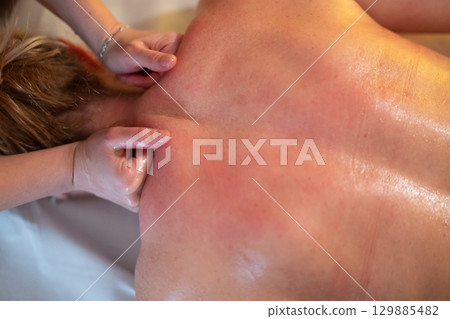 Relaxing Back Massage in a Spa Setting. A professional massage therapist in a blue uniform provides a relaxing back massage to a client lying on a massage table. Relaxing Back Massage in a Spa Setting. A professional massage therapist in a blue uniform provides a relaxing back massage to a client lying on a massage table. 129885482