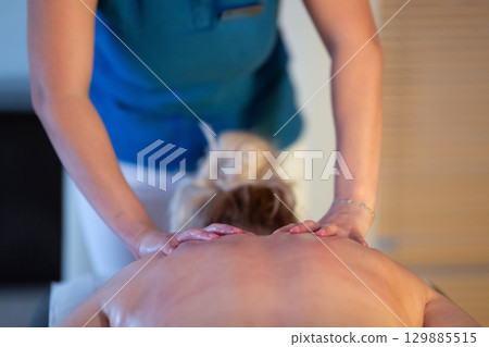 Relaxing Back Massage in a Spa Setting. A professional massage therapist in a blue uniform provides a relaxing back massage to a client lying on a massage table. 129885515