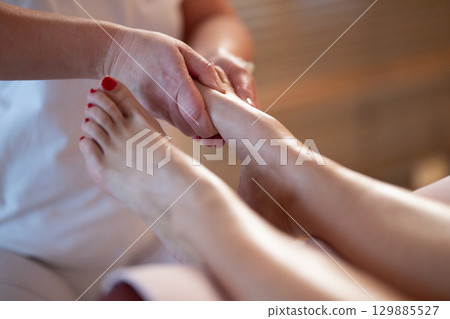 Foot Massage Therapy in a Relaxing Spa Setting. A professional massage therapist in a white uniform provides a soothing foot massage to a client in a calm and serene spa environment 129885527