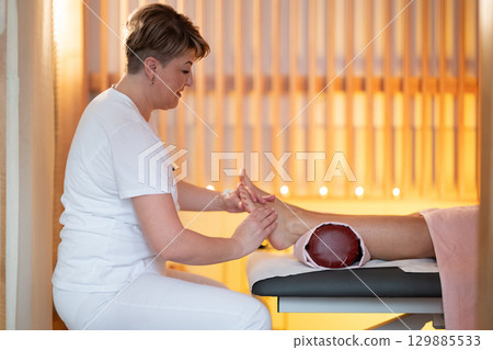 Foot Massage Therapy in a Relaxing Spa Setting. A professional massage therapist in a white uniform provides a soothing foot massage to a client in a calm and serene spa environment 129885533