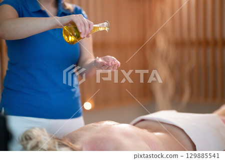 A professional massage therapist pours aromatic oil into their hands, preparing for a relaxing and rejuvenating massage session in a serene spa setting. 129885541