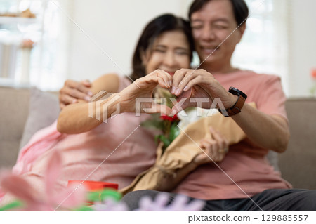 Romantic Couple Celebrating Valentine's Day with Heart Gesture and Flowers on Cozy Sofa Romantic Couple Celebrating Valentine's Day with Heart Gesture and Flowers on Cozy Sofa 129885557