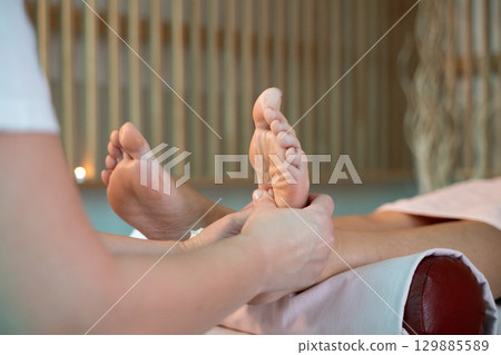 Foot Massage Therapy in a Relaxing Spa Setting. A professional massage therapist in a white uniform provides a soothing foot massage to a client in a calm and serene spa environment 129885589