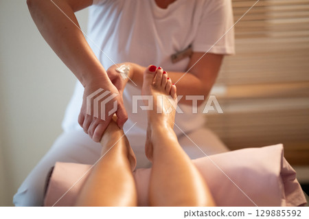 Foot Massage Therapy in a Relaxing Spa Setting. A professional massage therapist in a white uniform provides a soothing foot massage to a client in a calm and serene spa environment Foot Massage Therapy in a Relaxing Spa Setting. A professional massage therapist in a white uniform provides a soothing foot massage to a client in a calm and serene spa environment 129885592