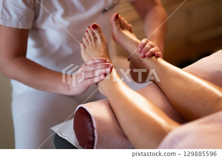 Foot Massage Therapy in a Relaxing Spa Setting. A professional massage therapist in a white uniform provides a soothing foot massage to a client in a calm and serene spa environment 129885596