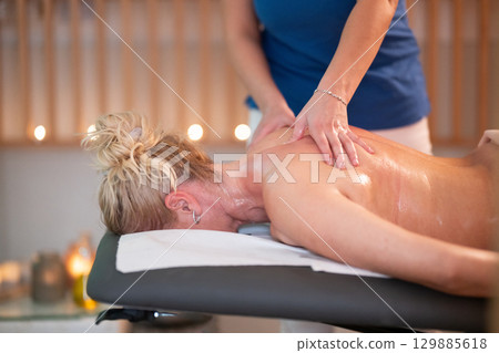 Relaxing Back Massage in a Spa Setting. A professional massage therapist in a blue uniform provides a relaxing back massage to a client lying on a massage table. 129885618
