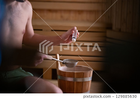 Essential Oils in Sauna. Close up of a mans hand holding essential oils inside a traditional wooden sauna with bucket and ladle. 129885630