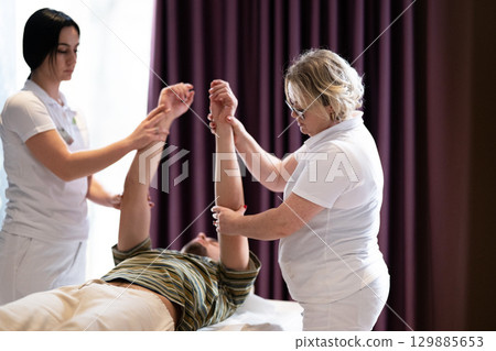 Two therapists assist a male patient in a physiotherapy session, focusing on arm rehabilitation exercises. 129885653
