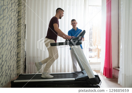 Physical Therapy on Treadmill.Man undergoing physical therapy with trainer assistance on a treadmill indoors. 129885659