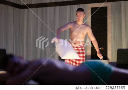 Hamam Attendant with Towel.Young male attendant preparing towel in traditional Turkish bath, standing near client. 129885665