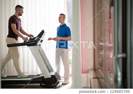 Physical Therapy on Treadmill.Man undergoing physical therapy with trainer assistance on a treadmill indoors. Physical Therapy on Treadmill.Man undergoing physical therapy with trainer assistance on a treadmill indoors. 129885700