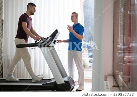 Physical Therapy on Treadmill.Man undergoing physical therapy with trainer assistance on a treadmill indoors. Physical Therapy on Treadmill.Man undergoing physical therapy with trainer assistance on a treadmill indoors. 129885738