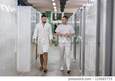 Medical Consultation in Spa Corridor.Two men discussing treatment options while walking through a clinical spa corridor, wearing white uniforms and bathrobe. 129885759