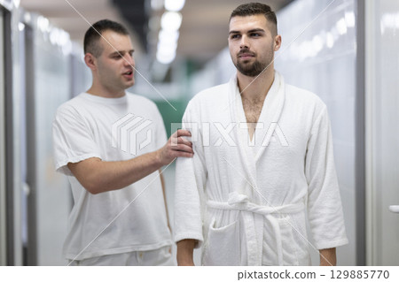 Medical Consultation in Spa Corridor.Two men discussing treatment options while walking through a clinical spa corridor, wearing white uniforms and bathrobe. Medical Consultation in Spa Corridor.Two men discussing treatment options while walking through a clinical spa corridor, wearing white uniforms and bathrobe. 129885770