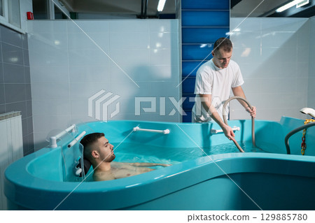 Therapeutic Water Bath.Man relaxing in hydrotherapy bath while therapist adjusts water settings. 129885780
