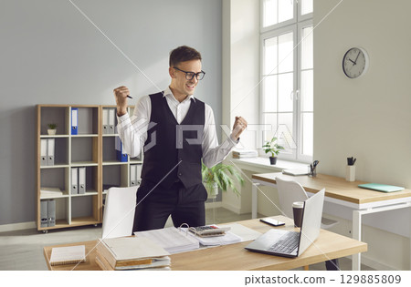 Happy man accountant working on laptop with documents in office rejoicing at success at work Happy man accountant working on laptop with documents in office rejoicing at success at work 129885809
