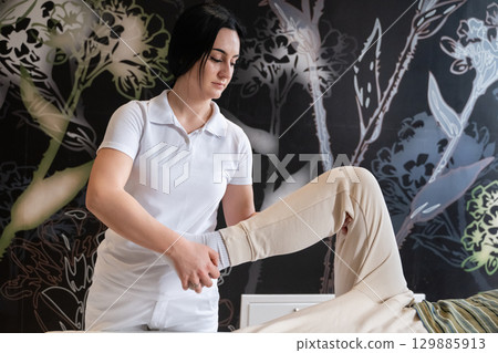 Physiotherapy Session with Blue Foam Roller.A therapist guiding a patient through leg exercises with the help of a blue foam roller in a therapy clinic. Physiotherapy Session with Blue Foam Roller.A therapist guiding a patient through leg exercises with the help of a blue foam roller in a therapy clinic. 129885913