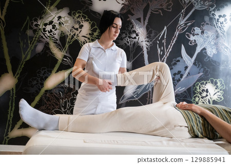 Physiotherapy Session with Blue Foam Roller.A therapist guiding a patient through leg exercises with the help of a blue foam roller in a therapy clinic. Physiotherapy Session with Blue Foam Roller.A therapist guiding a patient through leg exercises with the help of a blue foam roller in a therapy clinic. 129885941