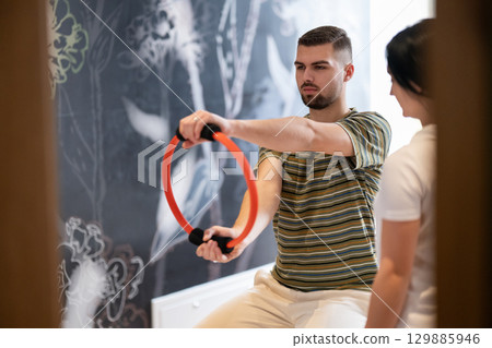 Physical Therapy Session with Resistance Ring.A male patient performing arm exercises using a resistance ring under supervision during a rehabilitation session. 129885946