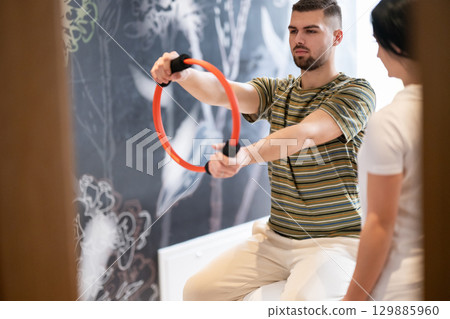 Physical Therapy Session with Resistance Ring.A male patient performing arm exercises using a resistance ring under supervision during a rehabilitation session. Physical Therapy Session with Resistance Ring.A male patient performing arm exercises using a resistance ring under supervision during a rehabilitation session. 129885960